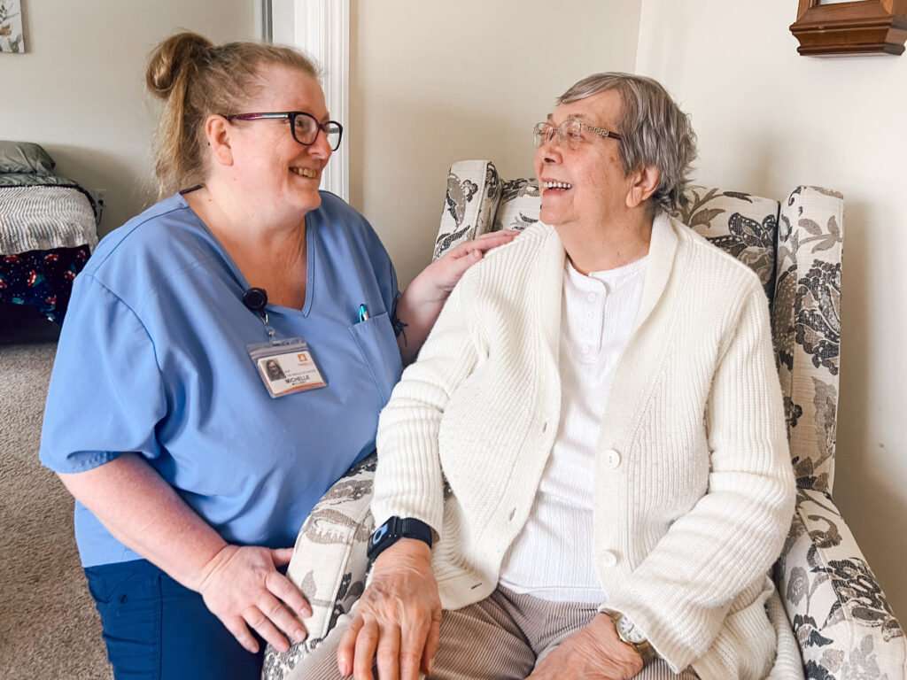 Caregiver in blue scrubs with a name badge chats warmly with an elderly woman in a beige cardigan, holding hands in a floral chair.