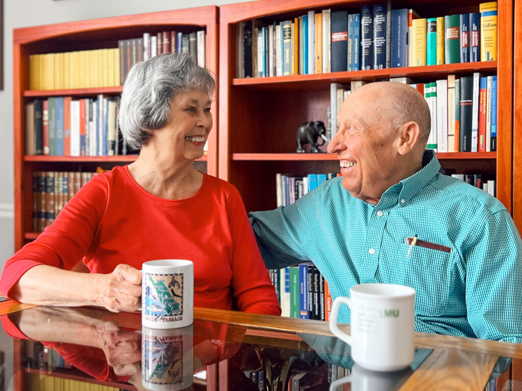 Two smiling seniors sit at a glass table with mugs, in front of red bookshelves filled with colorful books behind them.