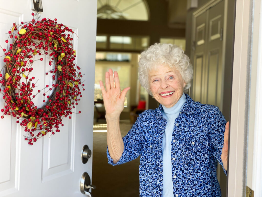 Smiling elderly woman standing in a doorway, waving hello, with a red berry wreath on a white door beside her.