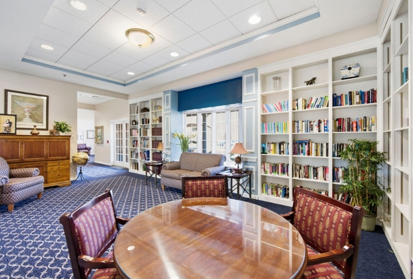 Public library lounge with a round wooden table and red upholstered chairs, surrounded by tall built-in bookshelves and a blue carpeted floor.