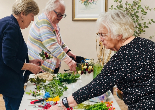 Three elderly people work together to arrange colorful flowers on a table indoors.