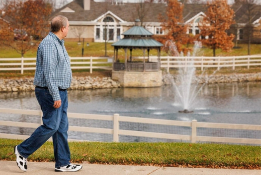Older man walking along a pond with a fountain, gazebo, and autumn trees in the background.