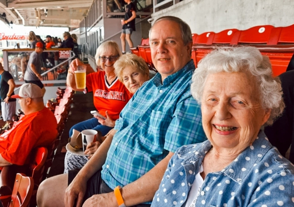 Smiling seniors seated in stadium bleachers at an outdoor event, some with drinks in hand.