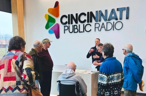 People gather around a reception counter under a Cincinnati Public Radio sign with a colorful logo, chatting and listening to staff.