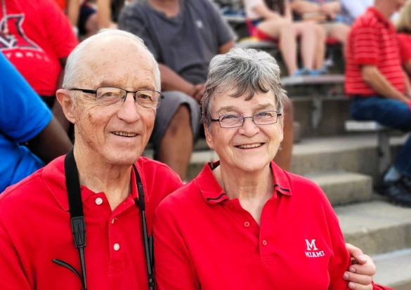 Smiling elderly man and woman in matching red polo shirts sit together at an outdoor stadium bleacher setting.