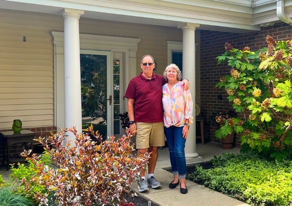 Couple stands on a front porch with white columns, smiling at the camera; man in a maroon polo and khaki shorts, woman in a colorful blouse and blue jeans.