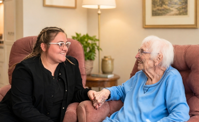 Caregiver and an elderly woman sharing a handshake and smiling in a cozy living room sitting on pink armchairs