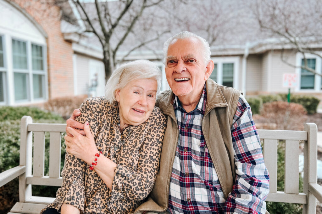 Two elderly friends hug and smile at a park bench, outdoors in front of a suburban home.