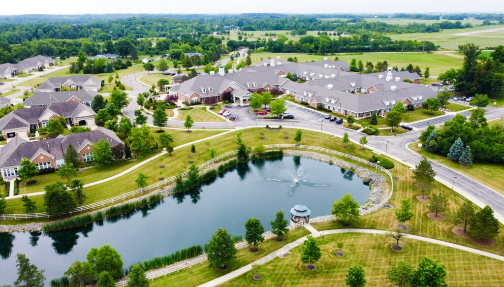 Aerial view of a suburban residential area around a pond with a fountain, walking paths, and green lawns with trees and houses nearby.
