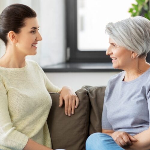 Two women sit on a sofa facing each other, smiling in a bright living room setting.3e0 They appear to be enjoying a friendly conversation on a couch with patterned pillows nearby.4843a8