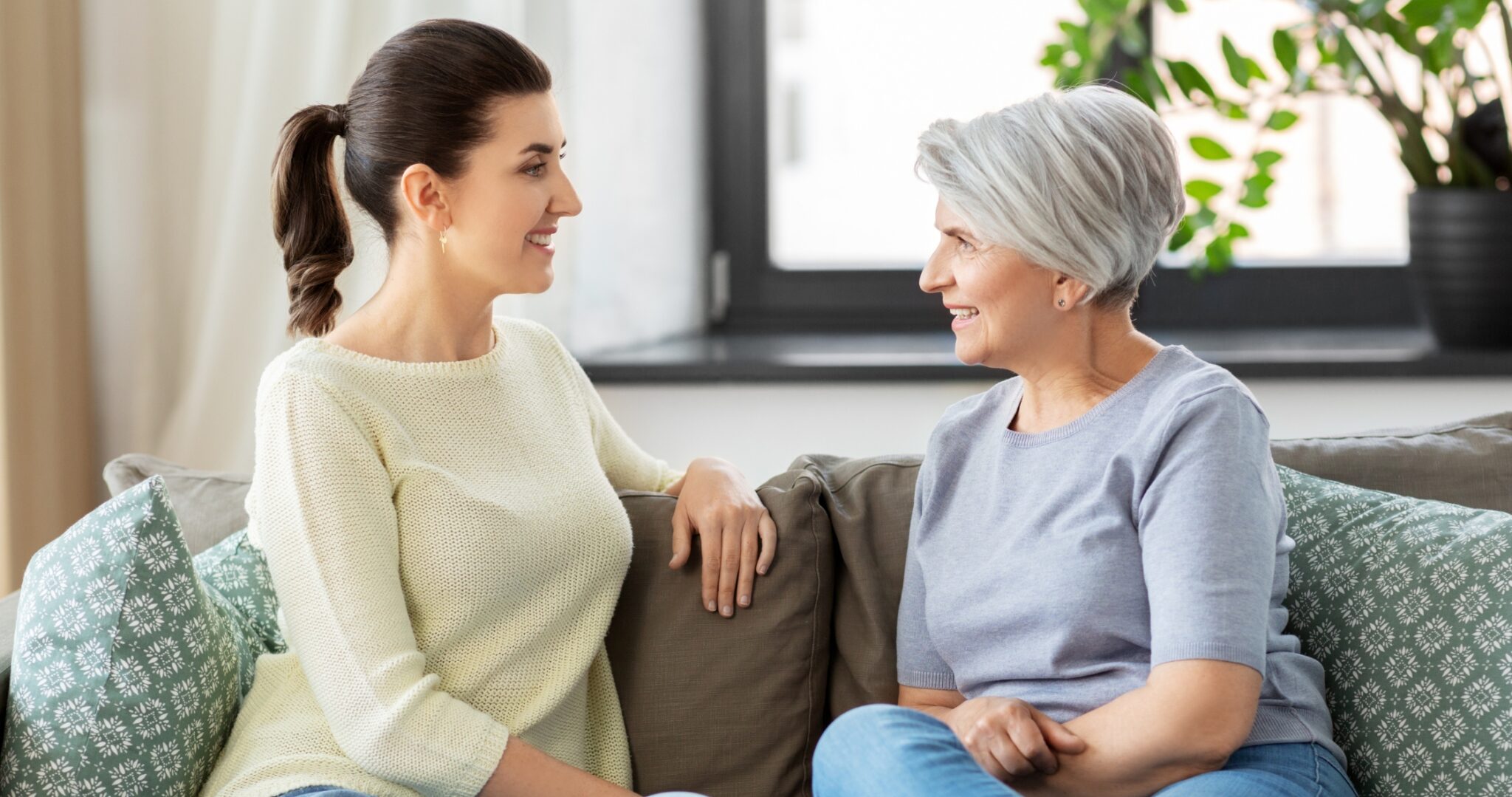 Two women sit on a sofa facing each other, smiling in a bright living room setting.3e0 They appear to be enjoying a friendly conversation on a couch with patterned pillows nearby.4843a8