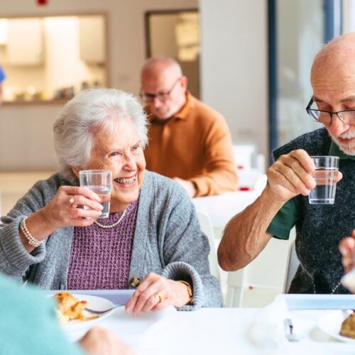 An elderly woman and man toast with water glasses at a group meal, smiling as they sit at a bright table with others in the background.