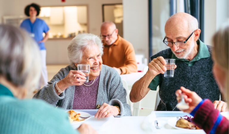 An elderly woman and man toast with water glasses at a group meal, smiling as they sit at a bright table with others in the background.