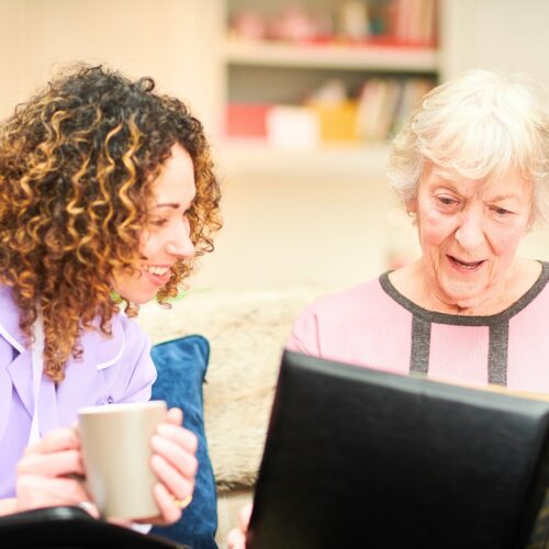 Caregiver in lavender scrubs sits with an elderly woman on a sofa, looking through a large photo album together, with mugs nearby.