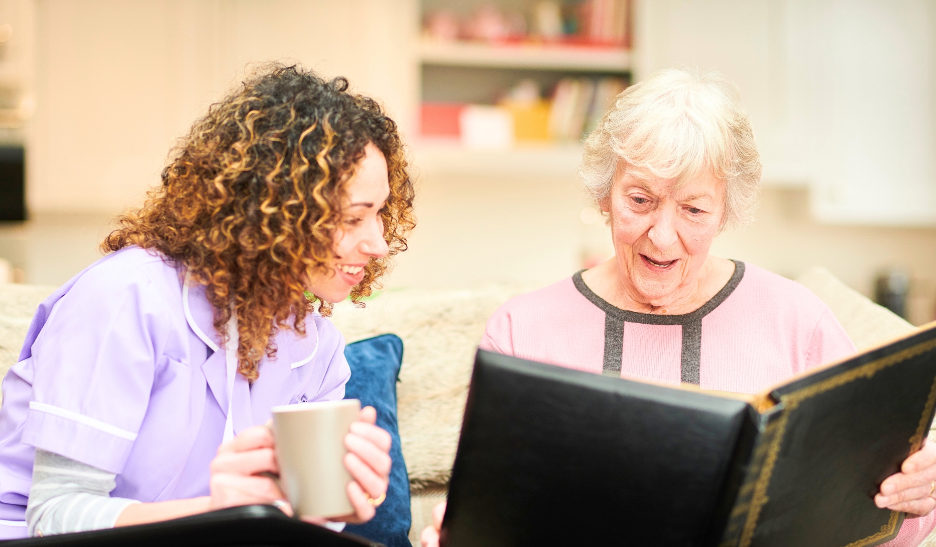 Caregiver in lavender scrubs sits with an elderly woman on a sofa, looking through a large photo album together, with mugs nearby.